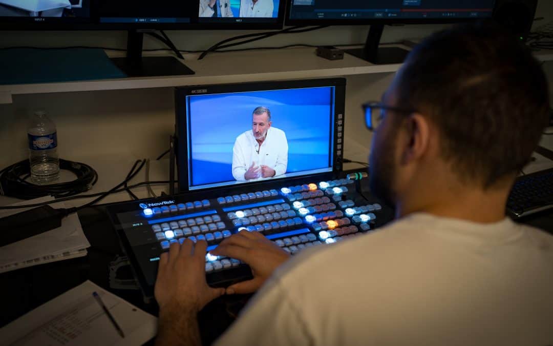TV studio control room. Operator at NewTek video switcher monitors a broadcast. Cristalina water bottle visible.